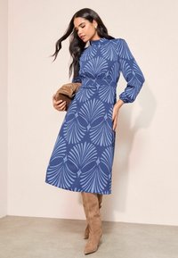 Zila midi kleita ar lapu rakstu, garām piedurknēm, augstu apkakli un jostu viduklī. Saskaņota ar brūnām augstām zābakiem.