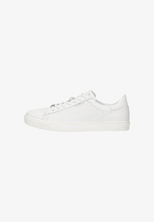 Personne portant un short noir et des baskets blanches basses debout sur un sol blanc.
