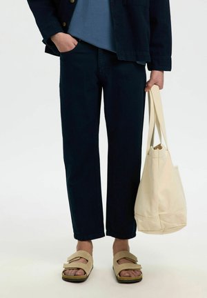 Personne portant un pantalon noir, des sandales beiges, tenant un sac cabas en toile beige, avec une main dans la poche et une chemise bleue partiellement visible.
