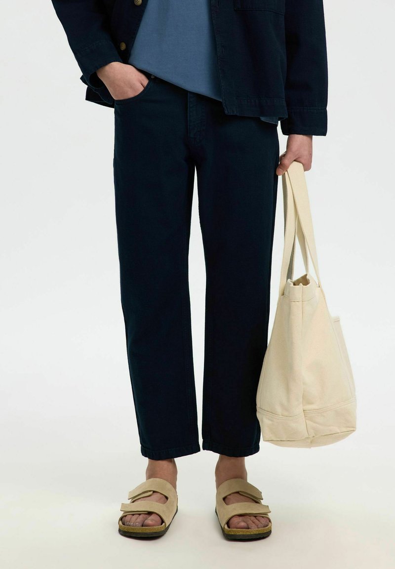 Personne portant un pantalon noir, des sandales beiges, tenant un sac cabas en toile beige, avec une main dans la poche et une chemise bleue partiellement visible.