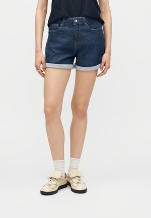 Femeie purtând pantaloni scurți din denim albastru închis, cu tiv rulodat, șosete albe, adidași bej și un top negru fără mâneci, stând pe un fundal simplu.