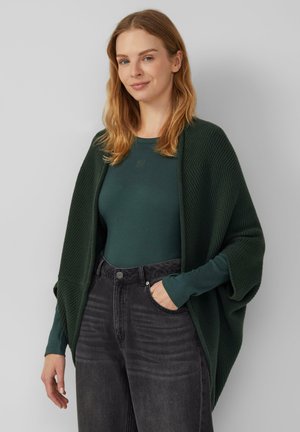 Donkergroene gebreide wikkelcardigan over een bijpassend langhemd met lange mouwen en zwarte jeans. Heeft losse mouwen en een textuurpatroon.