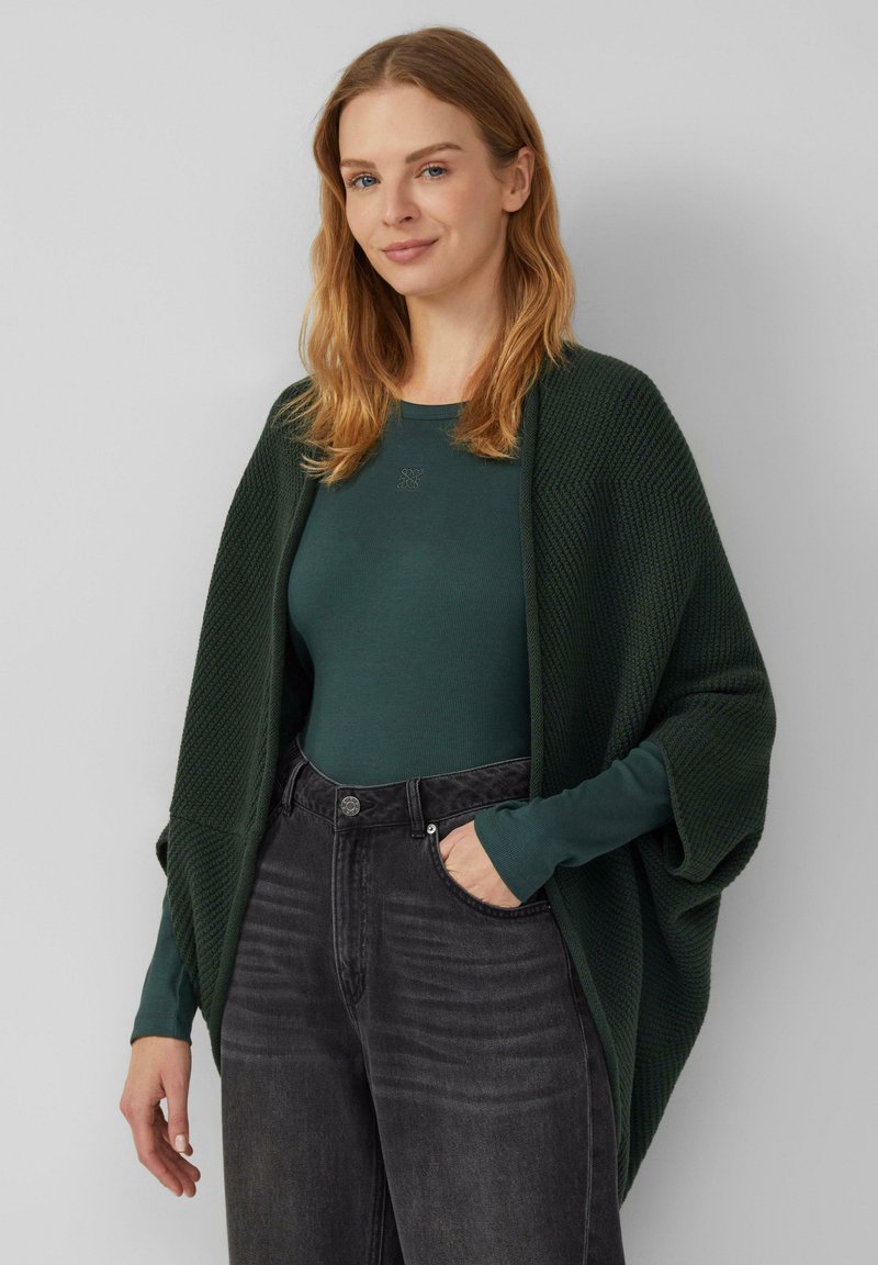 Dunkelgrüne gestrickte Wickeljacke, getragen über einem passenden Langarmoberteil und schwarzen Jeans. Verfügt über lockere Ärmel und ein strukturiertes Muster.