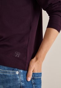 Burgundowa koszula z długim rękawem w luźnym kroju, z małym logo "Cozy" na dolnej krawędzi, zestawiona z niebieskimi dżinsami. Miękka, gładka tekstura.