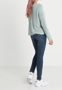 Světlý zelený pletený svetr, volnější střih, dlouhé rukávy. Kombinováno s tmavými úzkými džínami a bílými teniskami s černými detaily.