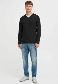 Black long-sleeve V-neck sweater paired with light blue denim jeans and gray sneakers, standing against a plain white background.