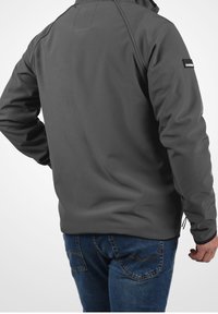 Grau weiche Softshell-Jacke mit hohem Kragen, Reißverschluss-Taschen und elastischen Bündchen. Auffälliges Logo am linken Ärmel. Kombiniert mit blauen Jeans.