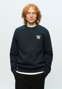 Navy sweatshirt with a round neckline and long sleeves, featuring a cream "W" logo on the chest, worn over a white shirt.