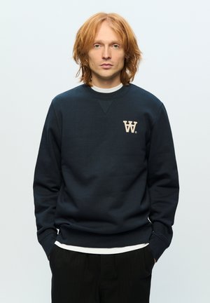 Námornícky sveter s okrúhlym výstrihom a dlhými rukávmi, s krémovým logom "W" na hrudi, nosený cez bielu košeľu.