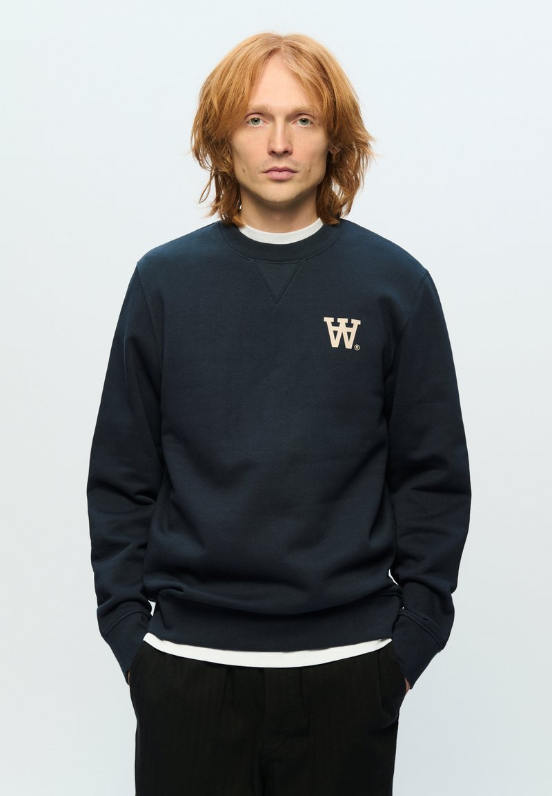 Navy sweatshirt with a round neckline and long sleeves, featuring a cream "W" logo on the chest, worn over a white shirt.