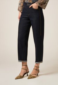 Vaqueros de mezclilla azul oscuro con dobladillos enrollados, que presentan costuras en dorado contrastante y un diseño clásico de cinco bolsillos. Combinados con tacones con un patrón.
