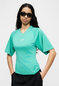 Γυναίκα με μακριά σκούρα μαλλιά που φοράει μαύρα γυαλιά ηλίου, μια τιρκουάζ αθλητική μπλούζα Puma και μαύρο παντελόνι με τα χέρια στις τσέπες, μπροστά σε λευκό φόντο.