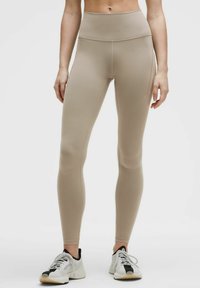 Leggings beige a vita alta realizzati in tessuto elasticizzato, con una texture liscia e un design aderente. Abbinati a scarpe da ginnastica bianche.