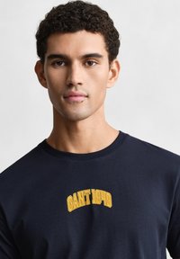 Camiseta de algodón azul marino con la inscripción "GANT 1949" en bordado amarillo en el pecho. Cuello redondo estándar, mangas cortas, textura suave.