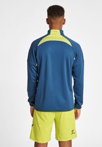 Maglia atletica a maniche lunghe in navy con tessuto testurizzato, dotata di accenti giallo brillante sulle spalle e sui polsini, indossata con pantaloni corti coordinati.