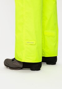 Pantaloni impermeabili giallo neon con una leggera texture, dotati di una patch con il logo "CEPLEX ACTIVE" e dettagli in velcro, abbinati a scarpe da ciclismo nere.