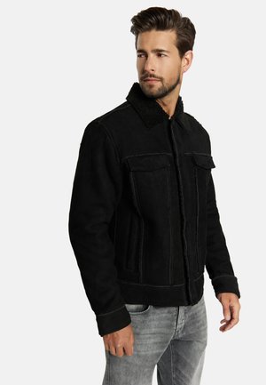 Mann mit kurzen Haaren und Bart, der eine schwarze Lammfelljacke und hellgraue Jeans trägt, schaut nach links vor einfarbigem Hintergrund.