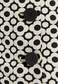 Svart och creamfärgad tyg med ett blommönster, som har stora svarta mittpunkter och mindre omgivande kronblad; två platta svarta knappar synliga.
