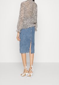 Blusa trasparente con motivo astratto marrone e crema, abbinata a una gonna in denim blu chiaro a lunghezza ginocchio con spacco posteriore e dettaglio dei bottoni.