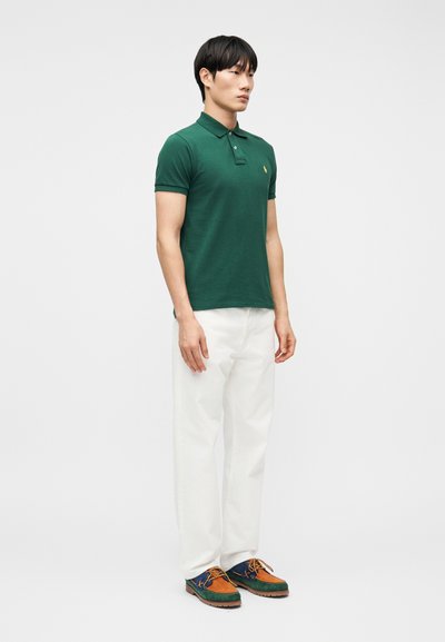 Άνδρας όρθιος, φορά πράσιν ο πόλο πουκάμισο, άσπρο παντελόνι και πολύχρωμα παπούτσια σε καφέ, μπλε και πράσινο χρώμα, σε απλό ανοιχτό φόντο.