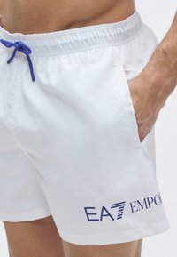 Fehér úszónadrág kék húzózsinóros derékpánttal, oldalzsebekkel és kék "EA7 EMPORIO" logóval a bal lábszáron.