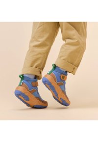 Sneaker blu e beige con tomaia in mesh testurizzato, fascia regolabile, linguetta verde sul tallone e suola in gomma audace, indossati con pantaloni beige arrotolati.