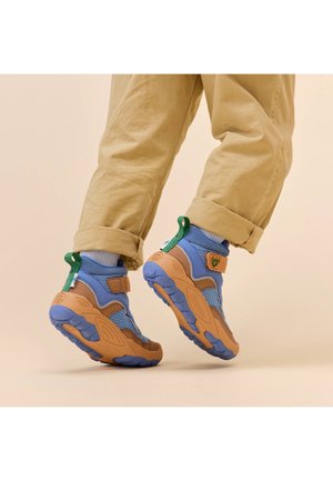Sneaker blu e beige con tomaia in mesh testurizzato, fascia regolabile, linguetta verde sul tallone e suola in gomma audace, indossati con pantaloni beige arrotolati.