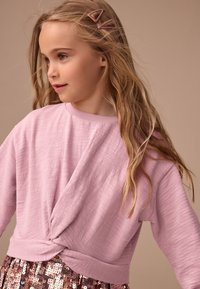 Világos rózsaszín hosszú ujjú felső, melynek derék részén csavaros dizájn található, texturált anyagból készült. Csillogó flitteres szoknyával párosítva.