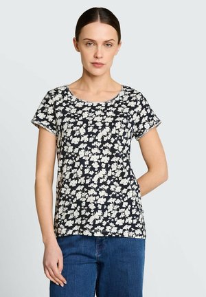 Kobieta o ciemnych włosach związanych z tyłu, ubrana w czarno-biały kwiecisty top z krótkim rękawem i niebieskie dżinsy, stojąca na jasnym tle.