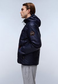 Giubbotto imbottito blu navy con cappuccio, design trapuntato, tasche con zip e patch con logo sulla manica sinistra; texture liscia e lucida.
