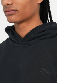 Črna bombažna kapuca z dvojno plastjo na pokrovčku, z diskretnim toniranim vezenjem logotipa na prsih. Gladka tekstura tkanine, sproščen kroj.