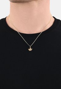 Gul halsband med fågelpendant, med fina kedjelänkar; bärs över en svart crewneck-tröja. Pendanten visar upp intrikata detaljer.