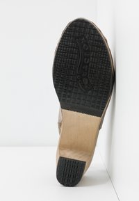 Semelle d’une chaussure beige avec une semelle en caoutchouc noir texturé au talon et à l’avant, montrant une bande de roulement à motif et un nom de marque partiellement visible.