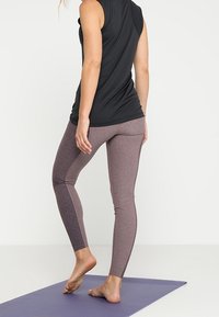 Musta ilmanhihaisa toppi ja mauve leggingsit, joissa on tiukka muotoilu. Leggingsit näyttävät kaksiväriseltä ja niissä on sileä tekstuuri. Seisoo joogamatolla.