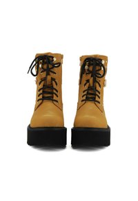 Un paio di stivali platform beige con lacci, suola spessa nera e occhielli in metallo decorativi, mostrati dal fronte su uno sfondo bianco.