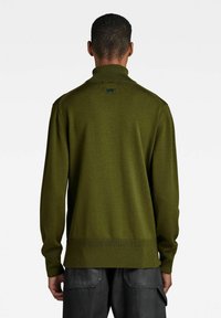 Olivgrüner Pullover mit hohem Kragen, geripptem Saum und Ärmeln sowie einem strukturierten Strickmuster. Dezente Markenkennzeichnung in der Nähe des Kragens.