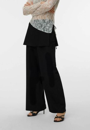 Person iført sorte vidde bukser med wrap-detalje, gennemsigtig hvid blondebluse med lange ærmer og sorte stroppesandaler med høje hæle.