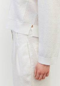 Lekki biały sweter dziany z bocznymi rozcięciami i kieszenią, zestawiony z białymi lnianymi spodniami z wysokim stanem i przednimi zakładkami.