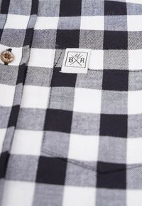 Rutig skjorta med knappar i svart och vitt tyg. Har en patchetikett med "BR" och en träknapp. Mjuk textur med en klassisk design.