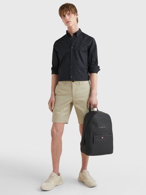 Schwarzes Hemd mit Knöpfen, beigefarbene Shorts, beigefarbene Sneakers und ein schwarzer Rucksack mit Struktur, einem vorderen Reißverschlussfach und Logodetail.