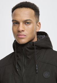 Schwarze Jacke mit Reißverschluss und verstellbarem Kapuze, mit einem runden Logo-Patch auf der Brust und einer glatten Textur.