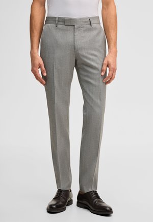 Uomo che indossa pantaloni grigi chiaro su misura, camicia bianca e scarpe eleganti in pelle nera, in piedi su uno sfondo semplice.