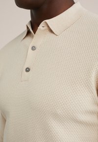Beige textured polo shirt with a woven pattern, featuring a collared neck and two dark grey buttons at the placket.