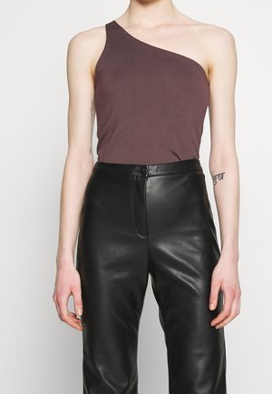 Henkilö, jolla on päällään hihaton, yhdellä olkapäällä ruskea toppi ja korkeavyötäröiset mustat nahkahousut, seisoo yksinkertaisen taustan edessä.