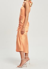 Robe midi en satin de couleur pêche douce avec fronces sur les côtés et bretelles en nœud ; portée avec des sandales blanches à talons ouverts. Texture lisse.