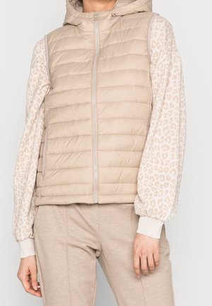 Jasnobeżowa pikowana kamizelka bez rękawów z przednim zamkiem błyskawicznym, noszona na jasnobeżowej bluzce z długim rękawem w delikatny lampartowy wzór oraz dopasowanych jasnobeżowych spodniach.