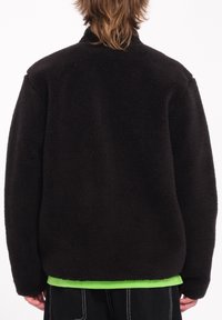 Zwarte fleece jacket met een gestructureerde oppervlakte, hoge kraag en verlaagde schouders. Gelaagd over een felgroen shirt met een rechte zoom.