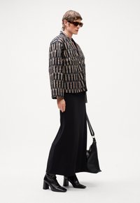 Jachetă cu model negru, căptușită, cu imprimeu geometric, asortată cu o rochie lungă neagră, cizme negre până la glezne și o geantă țesută. Ochelari de soare marron.