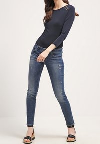 Žena oblečená v tmavě modrém dlouhém rukávu s detaily knoflíků, vycházkových úzkých džínách a černých klínových sandálech se zapínáním kolem kotníku.