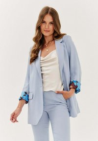 Světlomodrý přizpůsobený blazer s květinovým vzorem na manžetách, spárovaný s krémovým saténovým topem a odpovídajícími přiléhavými kalhotami. Sofistikované designové detaily.